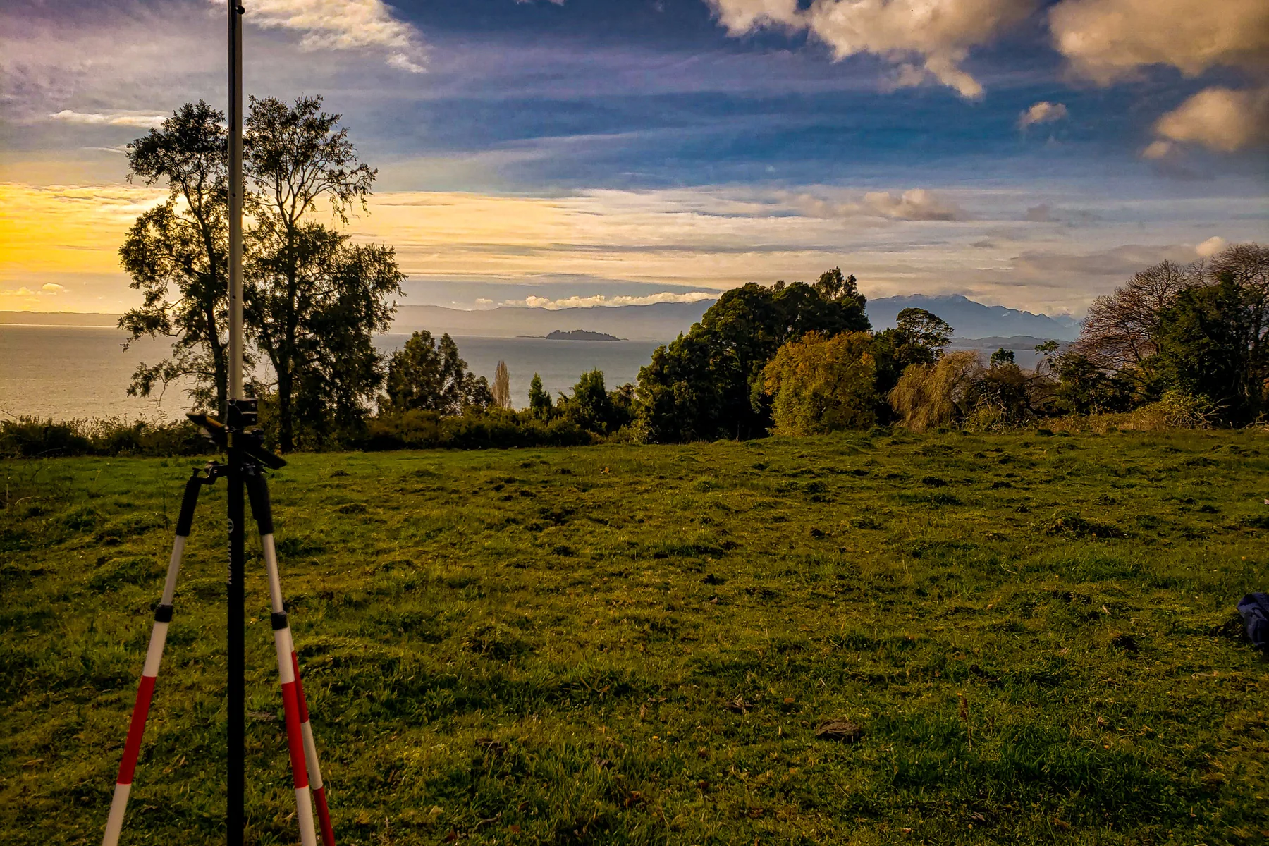 Jalones de topografía plantados en una pradera al atardecer, con el lago y los cerros al fondo.