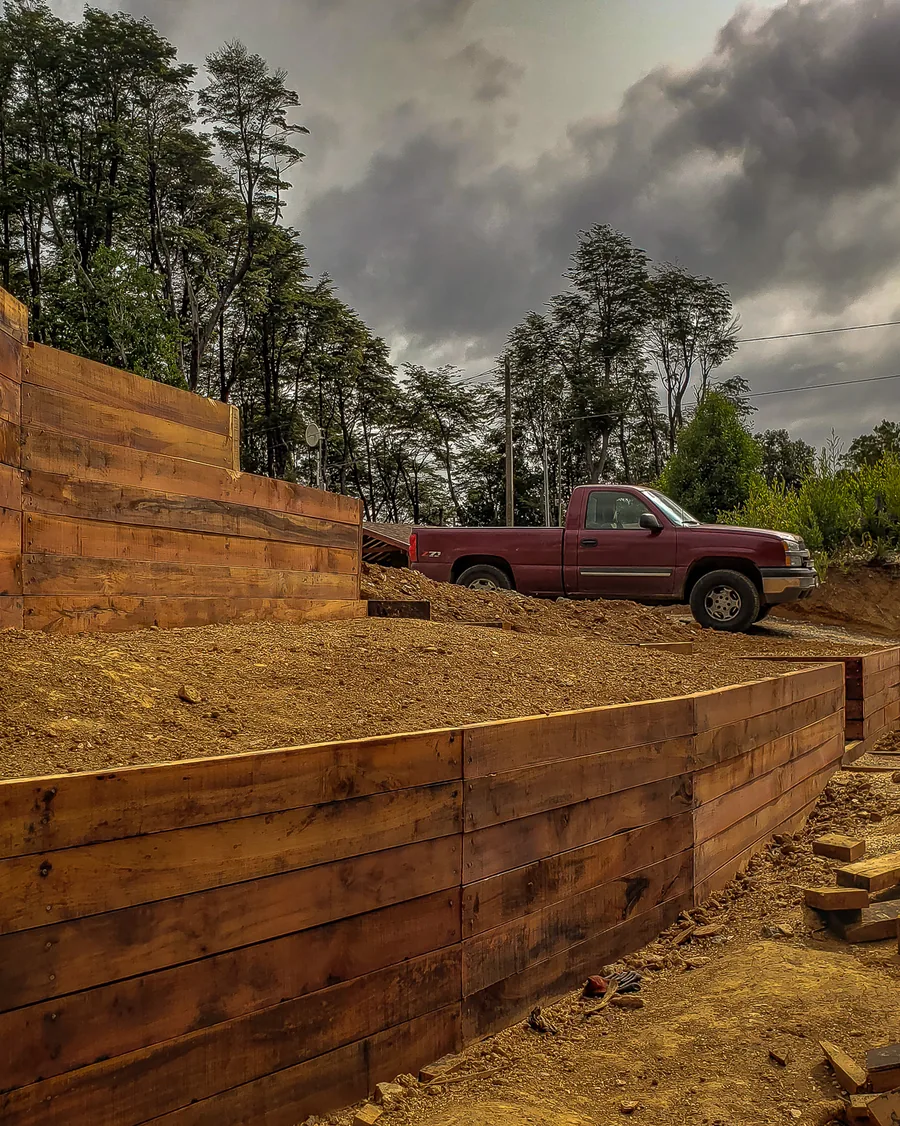 Muro de contención de madera en construcción, con la camioneta de faena bajo un cielo de tormenta.